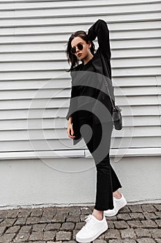Stylish young woman in fashionable youth black clothes in sunglasses in white trendy sneakers with leather handbag poses near