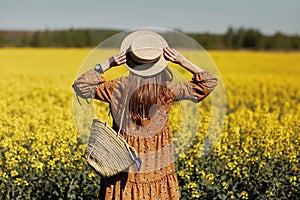 Stylish young woman in a field of yellow flowers. Girl in straw hat and in a floral dress and with a wicker bag