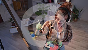 stylish young woman in glasses with dirty hands creates a picture using paints and brush, artist listens to music on