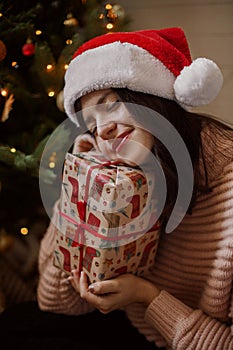 Stylish young woman holding christmas gift box and dreaming under christmas tree with lights in modern room. Happy girl in santa