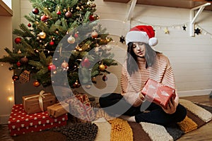 Stylish young woman holding red christmas gift box under christmas tree with lights in modern room. Happy girl in santa hat and