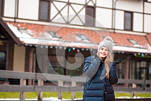 Stylish young woman in knitted hat and coat posing near the house. Space for text