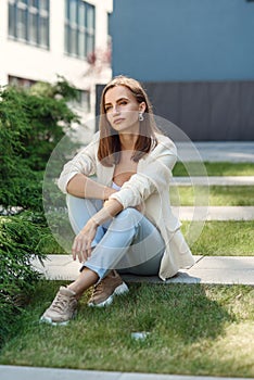 Stylish young woman at light casual outfit standing near the gray wall of the modern building.