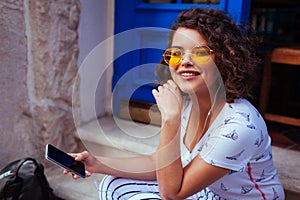 Stylish young woman listening to the music using smartphone. Outdoor portrait of fashionable girl wearing glasses