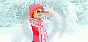 Stylish young woman looking away wearing sunglasses, colorful knitted hat and scarf in the park on snowy background