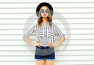 Stylish young woman model in black round hat, shorts, white striped shirt posing on white wall