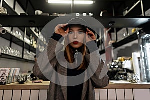 Stylish young woman with a pierced nose in a trendy black cap in a gray vintage checkered jacket with red lips posing at the bar.