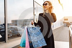 Stylish young woman in sunglasses and dress, with shopping bags, in the city against the background of a shopping center