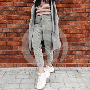 Stylish young woman in vintage pink sweatshirt in fashion jeans in gray long coat in fashionable sneakers stands near brick wall.