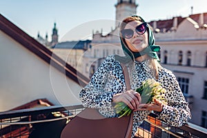 Stylish young woman wearing green retro shawl with sunglasses holding spring flowers in city. Classic vintage fashion