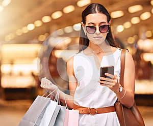 Stylish young woman wearing sunglasses using a cell phone while carrying bags during a shopping spree. Young brunette