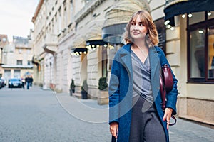 Stylish young woman wearing trendy outfit blue coat walking with purse outdoors. Spring fashion female accessories