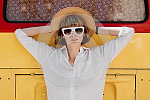 Stylish young woman in white sunglasses, a straw hat and white shirt poses next to the a bright red and yellow minivan