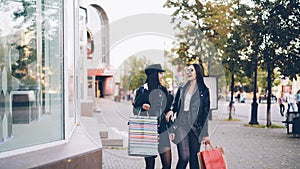 Stylish young women friends are walking along street holding paper bags with purchases and looking at shop windows