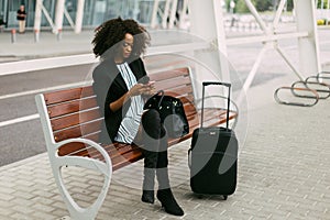 The stylishly dressed afro-a,erican traveler is sitting on the bench and chatting via the mobile phone.