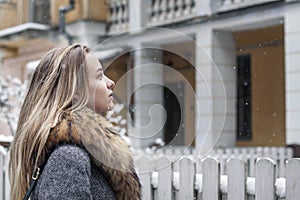 Stylishly dressed, beautiful girl in a fur coat, stands near the fence on the background of a residential building.