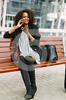 Stylishly dressed smiling afro-american traveler is talking via the mobile phone while sitting on the bench.