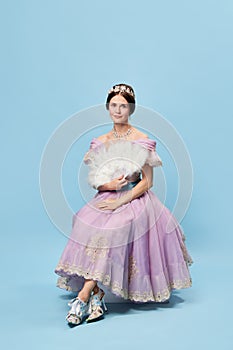 Stylishness. Beautiful charming girl in lilac color medieval dress as young queen or princess on blue background. Eras