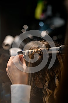 stylist makes curls hairstyle of hair with the curling iron in hairdresser salon, close up