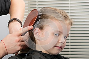 Stylist makes hair styling on the head with a comb for a little smiling girl