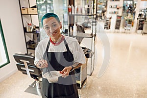 Stylist preparing a hair dye in a container and looks camera with smile. Hairdresser salon concept