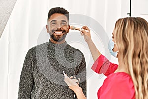 Stylist woman applying make up to handsome hispanic model at photo shoot backstage, getting ready for professional photoshoot