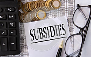 SUBSIDIES - word on a white piece of paper on the background of a calculator, pennies and glasses