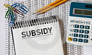 SUBSIDY - word in a notebook on the background of a table with numbers, calculator and pencils