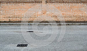 Suburban street background for copy space. Asphalt road with manholes in front of a sidewalk with weeds and a brickwall