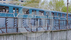 The subway train is moving behind a fence with barbed wire