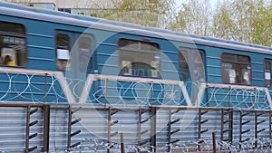 The subway train is moving behind a fence with barbed wire