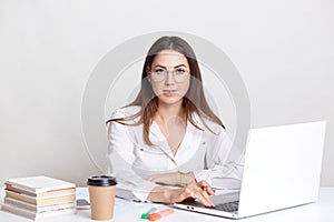 Succesful copywriter busy working with laptop computer, wears round glasses, drinks takeaway coffee, poses against white backgroun