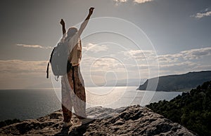 Success concept. Hiker with backpack standing on top of a mountain with raised hands and enjoying view