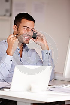 Success is mine. an smiling young man sitting at his desk and talking on his mobile phone.