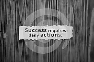 Success requires daily actions. Inspirational words on white torn paper note on wooden wall background in black and white.