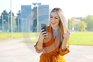 Success woman celebrating outdoor cheering and raising her fist up in exultation. Excited fashion woman watching her phone in the