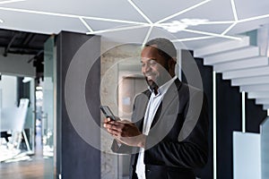 Successful african american businessman inside office standing by window, boss using phone, man typing message smiling