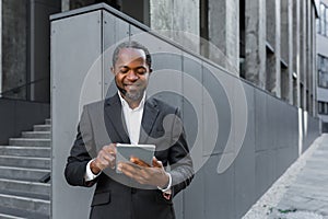 Successful african american man in business suit smiling and using tablet computer, businessman outside modern office