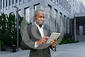 Successful african american man in business suit smiling and using tablet computer, businessman outside modern office