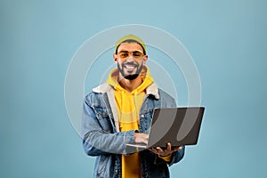 Successful arab freelancer guy using laptop computer and smiling to camera while standing over blue background