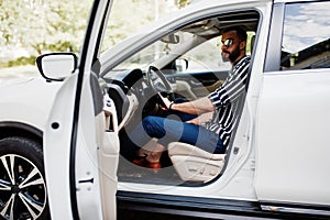 Successful arab man wear in striped shirt and sunglasses pose behind the wheel of  his white suv car. Stylish arabian men in