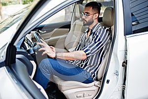 Successful arab man wear in striped shirt and sunglasses pose behind the wheel of  his white suv car. Stylish arabian men in