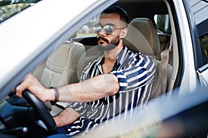 Successful arab man wear in striped shirt and sunglasses pose behind the wheel of  his white suv car. Stylish arabian men in