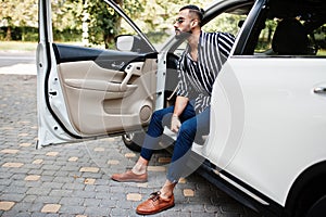 Successful arab man wear in striped shirt and sunglasses pose behind the wheel of  his white suv car. Stylish arabian men in