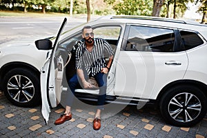 Successful arab man wear in striped shirt and sunglasses pose behind the wheel of  his white suv car. Stylish arabian men in