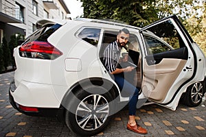 Successful arab man wear in striped shirt and sunglasses pose inside white suv car with laptop in hands
