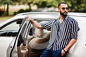 Successful arab man wear in striped shirt and sunglasses pose near his white suv car. Stylish arabian men in transport