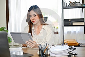 Successful Asian businesswoman at her desk, using tablet to recheck her financial data