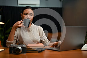 Successful Asian businesswoman sipping coffee while working on her project on laptop