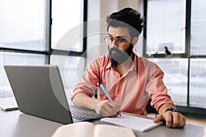 Successful bearded Indian businessman in glasses using laptop analyzing working project results, writing notes siting at